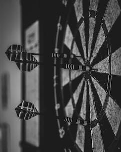 Close-up view of a dartboard with several darts embedded in it. The image is in black and white, highlighting the geometric patterns and textures of the board and darts.