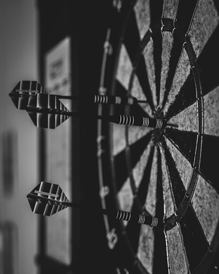 Close-up view of a dartboard with several darts embedded in it. The image is in black and white, highlighting the geometric patterns and textures of the board and darts.