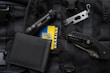 An assortment of tactical items lies on a black, molle backpack. The items include a black flashlight, a multi-tool, a folding knife, a black wallet, and a patch displaying the Ukrainian flag.