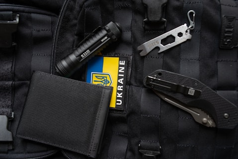 An assortment of tactical items lies on a black, molle backpack. The items include a black flashlight, a multi-tool, a folding knife, a black wallet, and a patch displaying the Ukrainian flag.