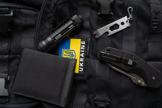 An assortment of tactical items lies on a black, molle backpack. The items include a black flashlight, a multi-tool, a folding knife, a black wallet, and a patch displaying the Ukrainian flag.