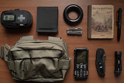 A neatly arranged collection of various items on a wooden surface, including a small first aid pouch, a black elastic band, a book resembling an old newspaper, a tactical pen, a wallet, a multitool, a flashlight, a folding knife, a rugged cellphone, and a green crossbody bag.