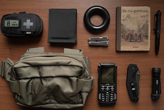 A neatly arranged collection of various items on a wooden surface, including a small first aid pouch, a black elastic band, a book resembling an old newspaper, a tactical pen, a wallet, a multitool, a flashlight, a folding knife, a rugged cellphone, and a green crossbody bag.