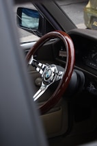 A vintage car interior showcasing a polished wooden steering wheel with a chrome center featuring a BMW logo. The view is from the passenger side, highlighting the retro dashboard and a glimpse of the side mirror.