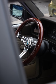 A vintage car interior showcasing a polished wooden steering wheel with a chrome center featuring a BMW logo. The view is from the passenger side, highlighting the retro dashboard and a glimpse of the side mirror.