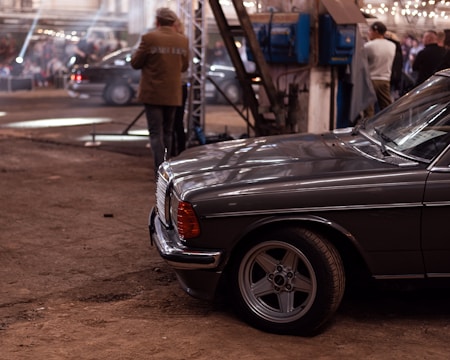 A vintage car with a classic design is parked in an industrial-style setting. The background features a large open area with people gathered, some standing and engaging in conversation. The environment has a warm tone, with exposed beams and string lights adding to the ambiance.