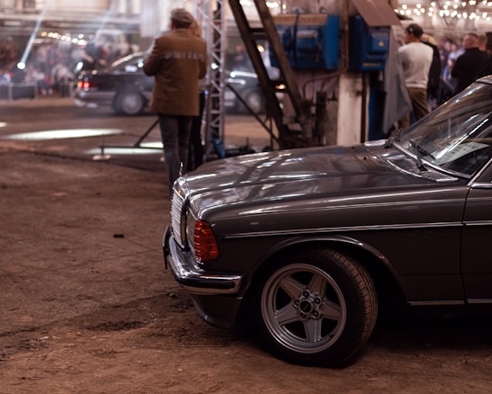 A vintage car with a classic design is parked in an industrial-style setting. The background features a large open area with people gathered, some standing and engaging in conversation. The environment has a warm tone, with exposed beams and string lights adding to the ambiance.