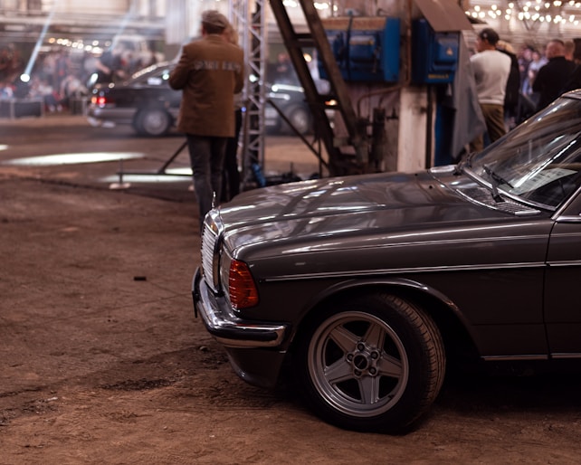 A vintage car with a classic design is parked in an industrial-style setting. The background features a large open area with people gathered, some standing and engaging in conversation. The environment has a warm tone, with exposed beams and string lights adding to the ambiance.