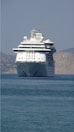 A large, white cruise ship is positioned prominently on the calm sea. The ship is facing the viewer, with its multiple decks and distinctive design clearly visible. In the background, hazy mountains provide a serene backdrop.