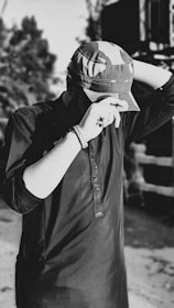A person wearing a dark kurta and a camouflage cap stands with one hand adjusting the cap and the other arm bent behind their head. The background is blurred with trees and a structure visible.