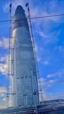 A tall, modern skyscraper under construction with a sleek, cylindrical design. It features glass windows and is flanked by cranes. The sky is clear with some scattered clouds, and some construction materials can be seen near the base.