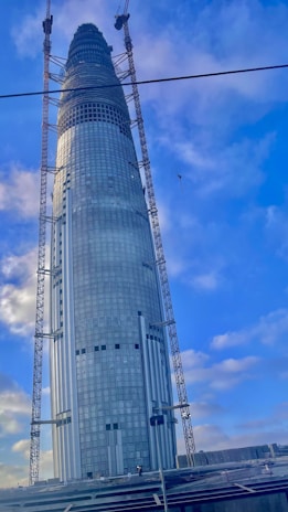 A tall, modern skyscraper under construction with a sleek, cylindrical design. It features glass windows and is flanked by cranes. The sky is clear with some scattered clouds, and some construction materials can be seen near the base.