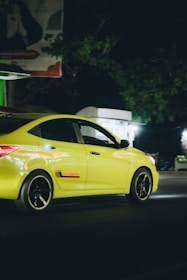 A bright yellow Hyundai car is driving on a dimly lit street. The car is moving at a noticeable speed, and its sleek design is highlighted by the streetlights. Shadows of surrounding trees and a faded billboard can be seen in the background.