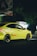 A bright yellow Hyundai car is driving on a dimly lit street. The car is moving at a noticeable speed, and its sleek design is highlighted by the streetlights. Shadows of surrounding trees and a faded billboard can be seen in the background.