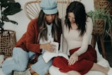 Two women are sitting on a cushioned surface, focusing on a book as one gestures toward its pages. The indoor setting includes woven furniture and green plants in the background. Both have long hair, with one wearing a cap and the other dressed in a long-sleeved top.