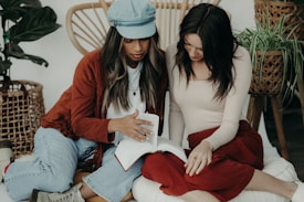 Two women are sitting on a cushioned surface, focusing on a book as one gestures toward its pages. The indoor setting includes woven furniture and green plants in the background. Both have long hair, with one wearing a cap and the other dressed in a long-sleeved top.
