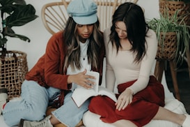 Two women are sitting on a cushioned surface, focusing on a book as one gestures toward its pages. The indoor setting includes woven furniture and green plants in the background. Both have long hair, with one wearing a cap and the other dressed in a long-sleeved top.