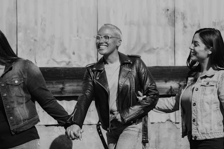 Three people holding hands, dressed casually in denim and leather jackets, stand in front of a textured wall. They appear joyful and are smiling, suggesting a sense of friendship and camaraderie.