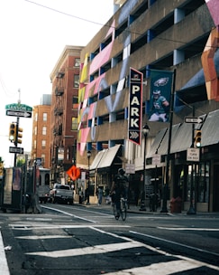 A bustling urban street scene featuring a multi-story parking garage adorned with vibrant, colorful murals. A person is riding a bicycle on the street, which is lined with various stores and businesses. Street signs, including ones indicating 'One Way,' 'No Turn on Red,' and street names like Sansom St., are visible. There are also traffic lights and vehicles present, and a banner displaying an image of a football player is attached to the parking structure.