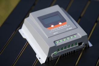 A close-up view of a solar charge controller labeled as Magic Series, featuring a digital display and multiple input/output connectors. The device is set against a metallic or reflective surface, suggesting an outdoor setup.