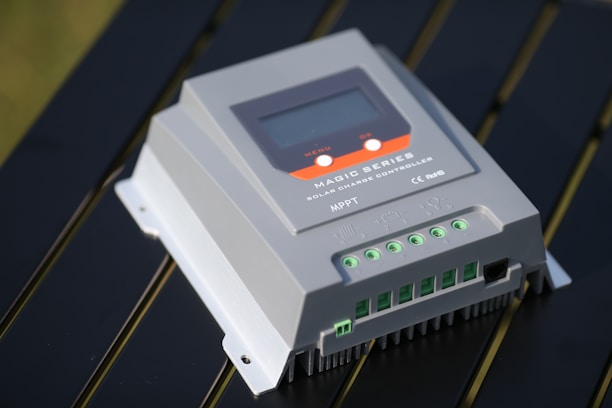 A close-up view of a solar charge controller labeled as Magic Series, featuring a digital display and multiple input/output connectors. The device is set against a metallic or reflective surface, suggesting an outdoor setup.