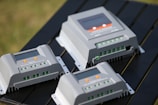 Several solar charge controllers are placed on a dark surface, featuring multiple green terminal connectors, an LED screen, and orange buttons. The controllers are positioned at different angles, highlighting their functionality and design.