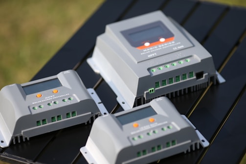 Several solar charge controllers are placed on a dark surface, featuring multiple green terminal connectors, an LED screen, and orange buttons. The controllers are positioned at different angles, highlighting their functionality and design.
