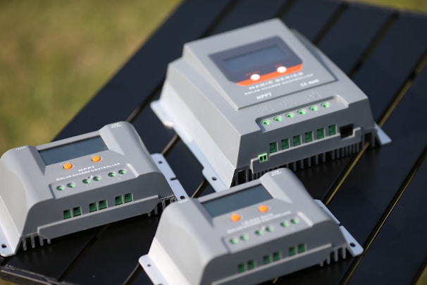 Several solar charge controllers are placed on a dark surface, featuring multiple green terminal connectors, an LED screen, and orange buttons. The controllers are positioned at different angles, highlighting their functionality and design.