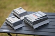 Three electronic devices, likely solar charge controllers, are placed on a black folding table. They are rectangular with a gray outer casing and various green and orange indicators on top. The background is a blur of greenery, indicating an outdoor setting.