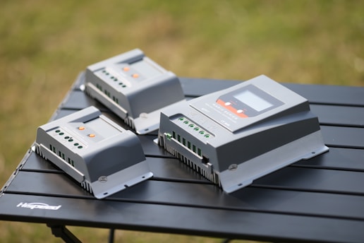 Three electronic devices, likely solar charge controllers, are placed on a black folding table. They are rectangular with a gray outer casing and various green and orange indicators on top. The background is a blur of greenery, indicating an outdoor setting.