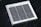 A small square solar panel with multiple black photovoltaic cells arranged in a grid pattern encased in a white frame. The panel is positioned on a dark, flat surface with a diagonal orientation.