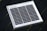 A small square solar panel with multiple black photovoltaic cells arranged in a grid pattern encased in a white frame. The panel is positioned on a dark, flat surface with a diagonal orientation.
