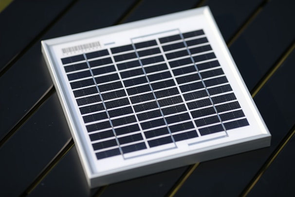 A small square solar panel with multiple black photovoltaic cells arranged in a grid pattern encased in a white frame. The panel is positioned on a dark, flat surface with a diagonal orientation.