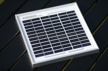 A small square solar panel with multiple black photovoltaic cells arranged in a grid pattern encased in a white frame. The panel is positioned on a dark, flat surface with a diagonal orientation.