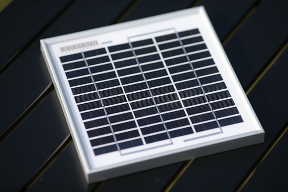A small square solar panel with multiple black photovoltaic cells arranged in a grid pattern encased in a white frame. The panel is positioned on a dark, flat surface with a diagonal orientation.