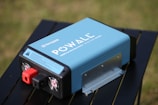 A blue portable power inverter with visible branding and specifications is placed on a black table outdoors. It has red and black connection ports and cooling fans on one end. The background features a blurred grassy area.