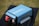 A blue portable power inverter with visible branding and specifications is placed on a black table outdoors. It has red and black connection ports and cooling fans on one end. The background features a blurred grassy area.