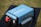 A blue portable power inverter with visible branding and specifications is placed on a black table outdoors. It has red and black connection ports and cooling fans on one end. The background features a blurred grassy area.