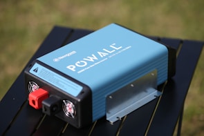 A blue portable power inverter with visible branding and specifications is placed on a black table outdoors. It has red and black connection ports and cooling fans on one end. The background features a blurred grassy area.