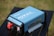 A blue portable power inverter with visible branding and specifications is placed on a black table outdoors. It has red and black connection ports and cooling fans on one end. The background features a blurred grassy area.