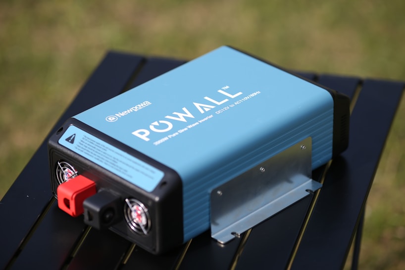A blue portable power inverter with visible branding and specifications is placed on a black table outdoors. It has red and black connection ports and cooling fans on one end. The background features a blurred grassy area.