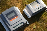 Two solar charge controllers labeled as 'Magic Series' are placed on a grassy surface. Both devices have digital displays, buttons, and terminals for electrical connections.