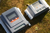 Two solar charge controllers labeled as 'Magic Series' are placed on a grassy surface. Both devices have digital displays, buttons, and terminals for electrical connections.