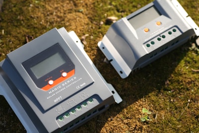 Two solar charge controllers labeled as 'Magic Series' are placed on a grassy surface. Both devices have digital displays, buttons, and terminals for electrical connections.