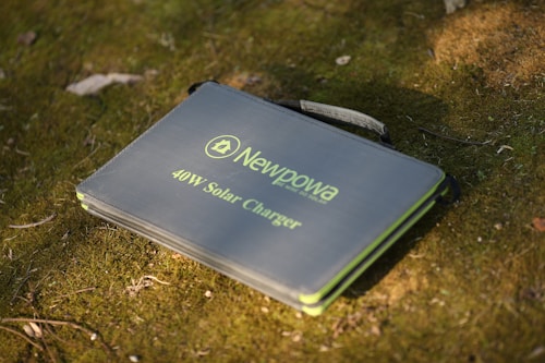 A portable solar charger with the brand name Newpowa and a 40W capacity is resting on grass or moss in a natural outdoor setting. The charger has a handle, suggesting it is designed for easy transport.