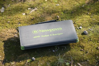 A folded Newpowa 40W solar charger is placed on a grassy surface, with a few scattered pebbles around it. The charger is in a case with a logo and text printed in green.