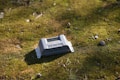 A small electronic device with buttons and connectors rests on a moss-covered ground. The surface is scattered with small stones and twigs, suggesting a natural outdoor setting.