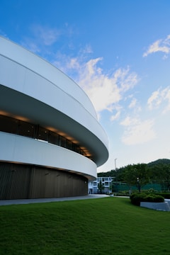 A modern, curved building with sleek architectural lines stands prominently against a bright blue sky. The structure is surrounded by a well-maintained green lawn and a few trees, with mountains visible in the background. Large windows are visible, suggesting a contemporary design with spacious interiors.
