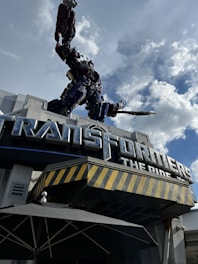 A large robot sculpture is positioned atop a building entrance, holding a weapon. The building features the sign 'TRANSFORMERS The Ride' in bold letters. The sky is filled with clouds, contributing to a dramatic scene.