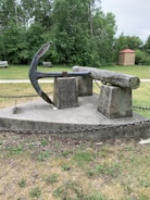 A large metal anchor and a thick wooden log are displayed on concrete pedestals, surrounded by a chain barrier. The scene is set in a grassy park area with lush green trees in the background. A small brick structure with a red roof stands nearby. The overall atmosphere is one of a historical or maritime display.